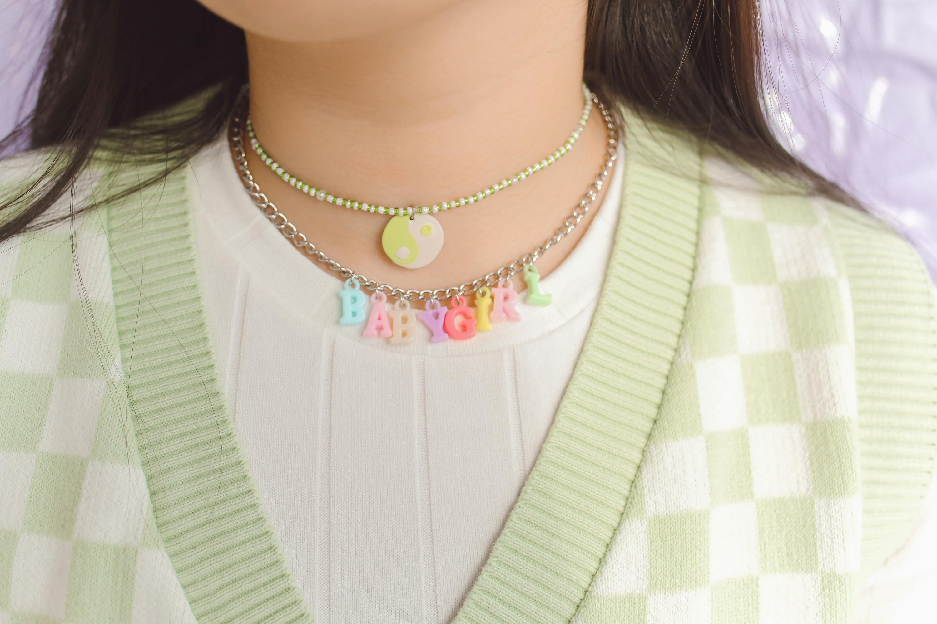 Close-up of layered pastel necklaces with beads spelling “BABYGIRL” over a white ribbed shirt and green checker cardigan.